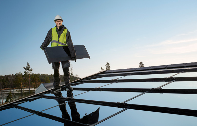 Man i arbetskläder står på tak med solpaneler och håller i en solpanel
